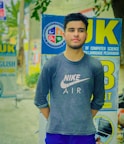A young man is standing outdoors wearing a dark Nike Air shirt with long sleeves. He has a short haircut and a well-groomed beard. Behind him are various signs for educational institutions, including one from the Department of Computer Science at an unspecified university. The background is slightly blurred, showing a mix of greenery and street signs.