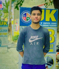 A young man is standing outdoors wearing a dark Nike Air shirt with long sleeves. He has a short haircut and a well-groomed beard. Behind him are various signs for educational institutions, including one from the Department of Computer Science at an unspecified university. The background is slightly blurred, showing a mix of greenery and street signs.