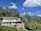 A modern two-story house with large windows surrounded by lush green trees and vegetation. The property includes a patio area with a satellite dish, and is situated in a picturesque landscape featuring mountains in the distance under a vibrant blue sky with scattered clouds.
