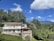 A modern two-story house with large windows surrounded by lush green trees and vegetation. The property includes a patio area with a satellite dish, and is situated in a picturesque landscape featuring mountains in the distance under a vibrant blue sky with scattered clouds.