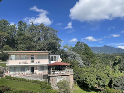 A modern two-story house with large windows surrounded by lush green trees and vegetation. The property includes a patio area with a satellite dish, and is situated in a picturesque landscape featuring mountains in the distance under a vibrant blue sky with scattered clouds.