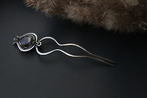 A delicate metal hairpin with an intricate design featuring a dark gemstone, placed on a dark surface. Soft, fluffy plant material fades into the background, adding a natural element.
