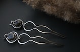 Two metallic hairpins designed with wavy, flowing shapes and black stone accents rest on a dark surface. The background includes a soft-textured, brownish, fluffy object that contrasts with the sleek, shiny material of the hairpins.