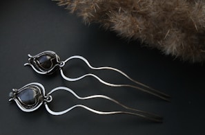 Two metallic hairpins designed with wavy, flowing shapes and black stone accents rest on a dark surface. The background includes a soft-textured, brownish, fluffy object that contrasts with the sleek, shiny material of the hairpins.