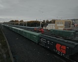 A long freight train with predominantly green cargo containers is seen on a railway track. The scene includes a red locomotive and a beige building nearby. Overhead power lines and autumnal trees in the background add to the industrial and serene atmosphere of the railway station.