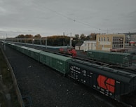 A long freight train with predominantly green cargo containers is seen on a railway track. The scene includes a red locomotive and a beige building nearby. Overhead power lines and autumnal trees in the background add to the industrial and serene atmosphere of the railway station.
