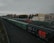 A long freight train with predominantly green cargo containers is seen on a railway track. The scene includes a red locomotive and a beige building nearby. Overhead power lines and autumnal trees in the background add to the industrial and serene atmosphere of the railway station.