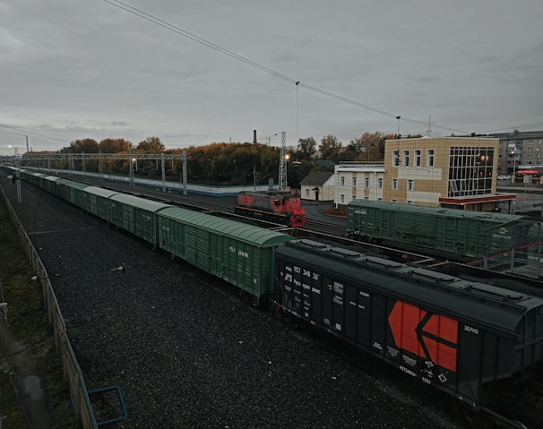 A long freight train with predominantly green cargo containers is seen on a railway track. The scene includes a red locomotive and a beige building nearby. Overhead power lines and autumnal trees in the background add to the industrial and serene atmosphere of the railway station.