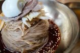 A close-up of a dish featuring Korean cold noodles neatly piled in a stainless steel bowl. The noodles are surrounded by a dark, spicy sauce with visible sesame seeds sprinkled on top. The dish is garnished with a slice of boiled egg, thinly sliced meat, and shredded vegetables.
