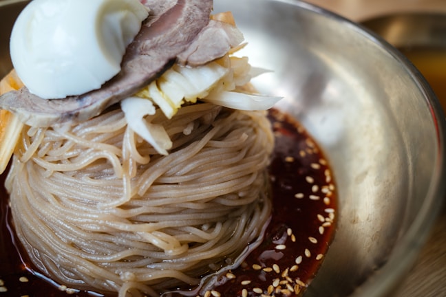 A close-up of a dish featuring Korean cold noodles neatly piled in a stainless steel bowl. The noodles are surrounded by a dark, spicy sauce with visible sesame seeds sprinkled on top. The dish is garnished with a slice of boiled egg, thinly sliced meat, and shredded vegetables.