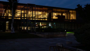 A modern glass building illuminated from the inside with a warm, yellow light. The upper floor of the building features a series of circular ceiling lights. The surroundings are dark, suggesting it is nighttime. There are some trees and greenery visible around the building.