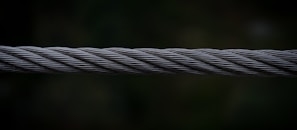 A close-up view of a twisted steel cable, with intricate strands interwoven tightly together. The cable is set against a dark, blurred background which emphasizes its metallic texture and structure.