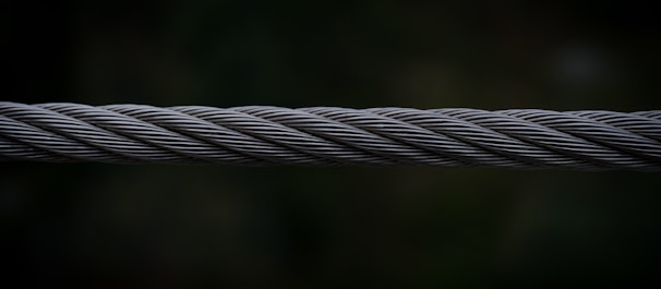 A close-up view of a twisted steel cable, with intricate strands interwoven tightly together. The cable is set against a dark, blurred background which emphasizes its metallic texture and structure.