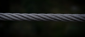 A close-up view of a twisted steel cable, with intricate strands interwoven tightly together. The cable is set against a dark, blurred background which emphasizes its metallic texture and structure.