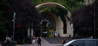 An arched entrance surrounded by lush greenery, with people walking and sitting around. The entrance has large windows and is topped with greenery, suggesting a blend of natural and architectural elements. The area is busy, with people engaged in various activities such as walking, sitting, and interacting with each other.