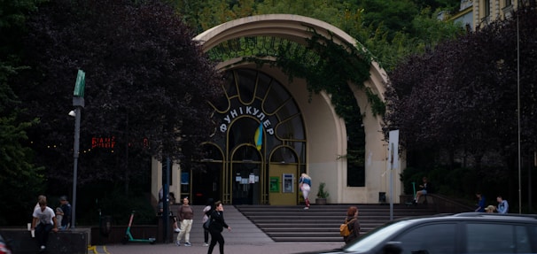 An arched entrance surrounded by lush greenery, with people walking and sitting around. The entrance has large windows and is topped with greenery, suggesting a blend of natural and architectural elements. The area is busy, with people engaged in various activities such as walking, sitting, and interacting with each other.