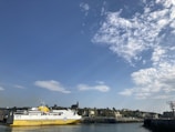 A large yellow and white ferry labeled 'Transmanche Ferries' is docked at a harbor. The background features a coastline with rocky cliffs and green vegetation. Above, there is a bright blue sky with scattered fluffy white clouds.