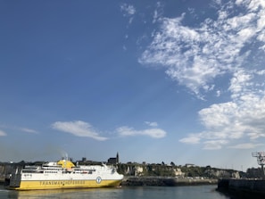 A large yellow and white ferry labeled 'Transmanche Ferries' is docked at a harbor. The background features a coastline with rocky cliffs and green vegetation. Above, there is a bright blue sky with scattered fluffy white clouds.