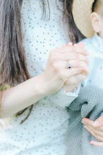 A close-up view of a person wearing a patterned shirt holding hands with a child. The child is wearing a straw hat and a light-colored shirt with small patterns, and is wrapped in a gray knitted blanket.