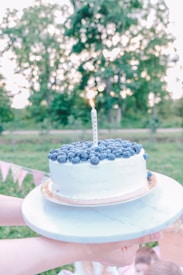 A round cake topped with fresh blueberries and a single lit candle is being held on a marble platter. In the background, there is a lush green outdoor setting with blurred trees and hanging bunting flags, indicating a festive atmosphere.