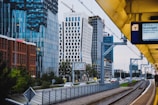 A modern urban scene featuring a cityscape with tall, contemporary office buildings characterized by glass facades and unique architectural designs. A train platform runs parallel to a road with passing cars, and a digital sign showing train information is visible. Overhead, there are construction cranes indicating ongoing development.