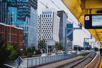 A modern urban scene featuring a cityscape with tall, contemporary office buildings characterized by glass facades and unique architectural designs. A train platform runs parallel to a road with passing cars, and a digital sign showing train information is visible. Overhead, there are construction cranes indicating ongoing development.