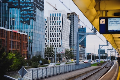 A modern urban scene featuring a cityscape with tall, contemporary office buildings characterized by glass facades and unique architectural designs. A train platform runs parallel to a road with passing cars, and a digital sign showing train information is visible. Overhead, there are construction cranes indicating ongoing development.
