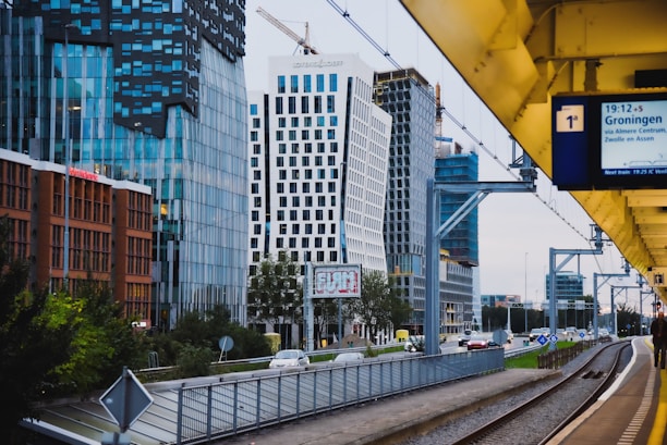 A modern urban scene featuring a cityscape with tall, contemporary office buildings characterized by glass facades and unique architectural designs. A train platform runs parallel to a road with passing cars, and a digital sign showing train information is visible. Overhead, there are construction cranes indicating ongoing development.
