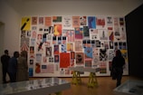 A gallery wall displaying a collection of vintage posters with various political and social messages in different colors and designs. Several people are observing the artwork, and the room features minimalist interiors with wooden floors and display tables.