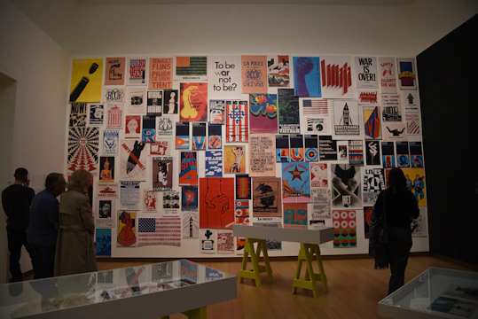 A gallery wall displaying a collection of vintage posters with various political and social messages in different colors and designs. Several people are observing the artwork, and the room features minimalist interiors with wooden floors and display tables.