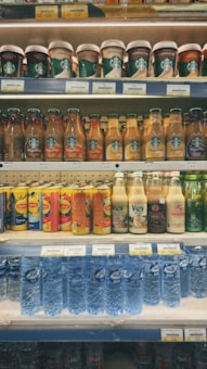 Various beverages are neatly arranged on multiple shelves in a supermarket. The top shelf contains cups of Starbucks beverages with visible labels. Below, rows of bottled drinks, including coffee, tea, and flavored water, are organized by brand. In the middle, there are fruit juices and plant-based drinks. The bottom shelf displays bottled mineral water.