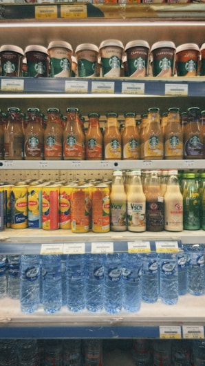 Various beverages are neatly arranged on multiple shelves in a supermarket. The top shelf contains cups of Starbucks beverages with visible labels. Below, rows of bottled drinks, including coffee, tea, and flavored water, are organized by brand. In the middle, there are fruit juices and plant-based drinks. The bottom shelf displays bottled mineral water.