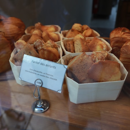 Several wooden baskets filled with assorted baked goods such as muffins and brownies are displayed. A small sign in French indicates the contents and price, mentioning chouquettes, financiers, madeleines, and chocolate cookies. The baked goods are golden brown and neatly arranged.