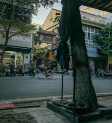A bustling urban street with a variety of elements, including a hair salon with a neon sign, motorcycles parked on the sidewalk, and a person riding a bicycle. The street is lined with buildings that have a mix of modern and older architectural styles. Trees provide some greenery and shade.