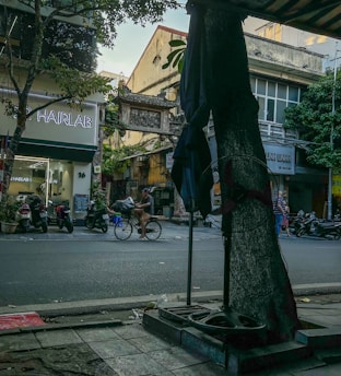 A bustling urban street with a variety of elements, including a hair salon with a neon sign, motorcycles parked on the sidewalk, and a person riding a bicycle. The street is lined with buildings that have a mix of modern and older architectural styles. Trees provide some greenery and shade.