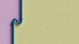 A blue pipe with an artistic bend is mounted on a two-tone wall. The wall features a lavender color on the left side and a light green color on the right. The pipe is secured with brackets and stands out against the muted background.