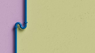 A blue pipe with an artistic bend is mounted on a two-tone wall. The wall features a lavender color on the left side and a light green color on the right. The pipe is secured with brackets and stands out against the muted background.