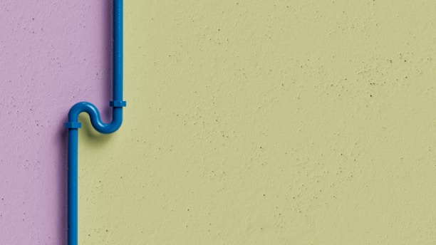 A blue pipe with an artistic bend is mounted on a two-tone wall. The wall features a lavender color on the left side and a light green color on the right. The pipe is secured with brackets and stands out against the muted background.