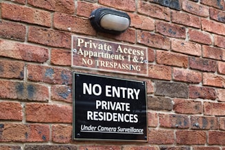 A brick wall with two signs posted on it. The upper sign reads 'Private Access, Apartments 1 & 2, No Trespassing' and the lower sign reads 'No Entry, Private Residences, Under Camera Surveillance'. There is also a light fixture above the signs.