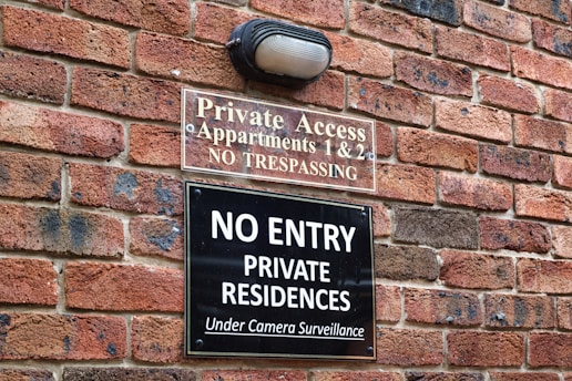 A brick wall with two signs posted on it. The upper sign reads 'Private Access, Apartments 1 & 2, No Trespassing' and the lower sign reads 'No Entry, Private Residences, Under Camera Surveillance'. There is also a light fixture above the signs.