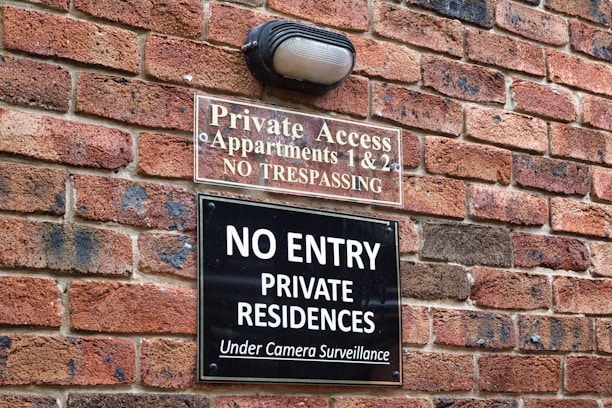 A brick wall with two signs posted on it. The upper sign reads 'Private Access, Apartments 1 & 2, No Trespassing' and the lower sign reads 'No Entry, Private Residences, Under Camera Surveillance'. There is also a light fixture above the signs.
