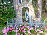 A serene statue of a religious figure is encased in a stone alcove amidst a forest setting. Pink flowers bloom vibrantly in front of the statue, and a metal fence surrounds the area, adding a sense of protection and reverence.