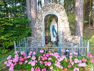 A serene statue of a religious figure is encased in a stone alcove amidst a forest setting. Pink flowers bloom vibrantly in front of the statue, and a metal fence surrounds the area, adding a sense of protection and reverence.