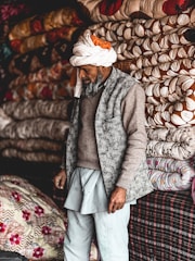 A man wearing a turban and traditional clothing stands next to a stack of intricately patterned quilts. The background is filled with colorful, neatly folded textiles in various designs, creating a vibrant and textured display.