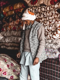 A man wearing a turban and traditional clothing stands next to a stack of intricately patterned quilts. The background is filled with colorful, neatly folded textiles in various designs, creating a vibrant and textured display.