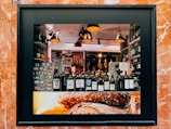A storefront display showcasing a variety of wine bottles arranged neatly on a shelf. The background features hanging cured meats and various food products. Warm, ambient lighting creates a cozy atmosphere, with modern pendant lamps adding to the aesthetic. The frame of the display is surrounded by reddish-brown marble tiles, and the lower part of the display features an image of rustic bread.