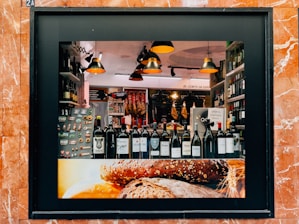 A storefront display showcasing a variety of wine bottles arranged neatly on a shelf. The background features hanging cured meats and various food products. Warm, ambient lighting creates a cozy atmosphere, with modern pendant lamps adding to the aesthetic. The frame of the display is surrounded by reddish-brown marble tiles, and the lower part of the display features an image of rustic bread.