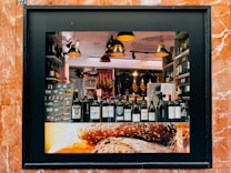 A storefront display showcasing a variety of wine bottles arranged neatly on a shelf. The background features hanging cured meats and various food products. Warm, ambient lighting creates a cozy atmosphere, with modern pendant lamps adding to the aesthetic. The frame of the display is surrounded by reddish-brown marble tiles, and the lower part of the display features an image of rustic bread.