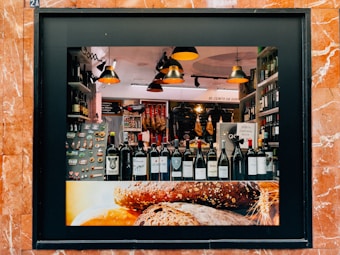 A storefront display showcasing a variety of wine bottles arranged neatly on a shelf. The background features hanging cured meats and various food products. Warm, ambient lighting creates a cozy atmosphere, with modern pendant lamps adding to the aesthetic. The frame of the display is surrounded by reddish-brown marble tiles, and the lower part of the display features an image of rustic bread.