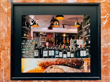 A storefront display showcasing a variety of wine bottles arranged neatly on a shelf. The background features hanging cured meats and various food products. Warm, ambient lighting creates a cozy atmosphere, with modern pendant lamps adding to the aesthetic. The frame of the display is surrounded by reddish-brown marble tiles, and the lower part of the display features an image of rustic bread.